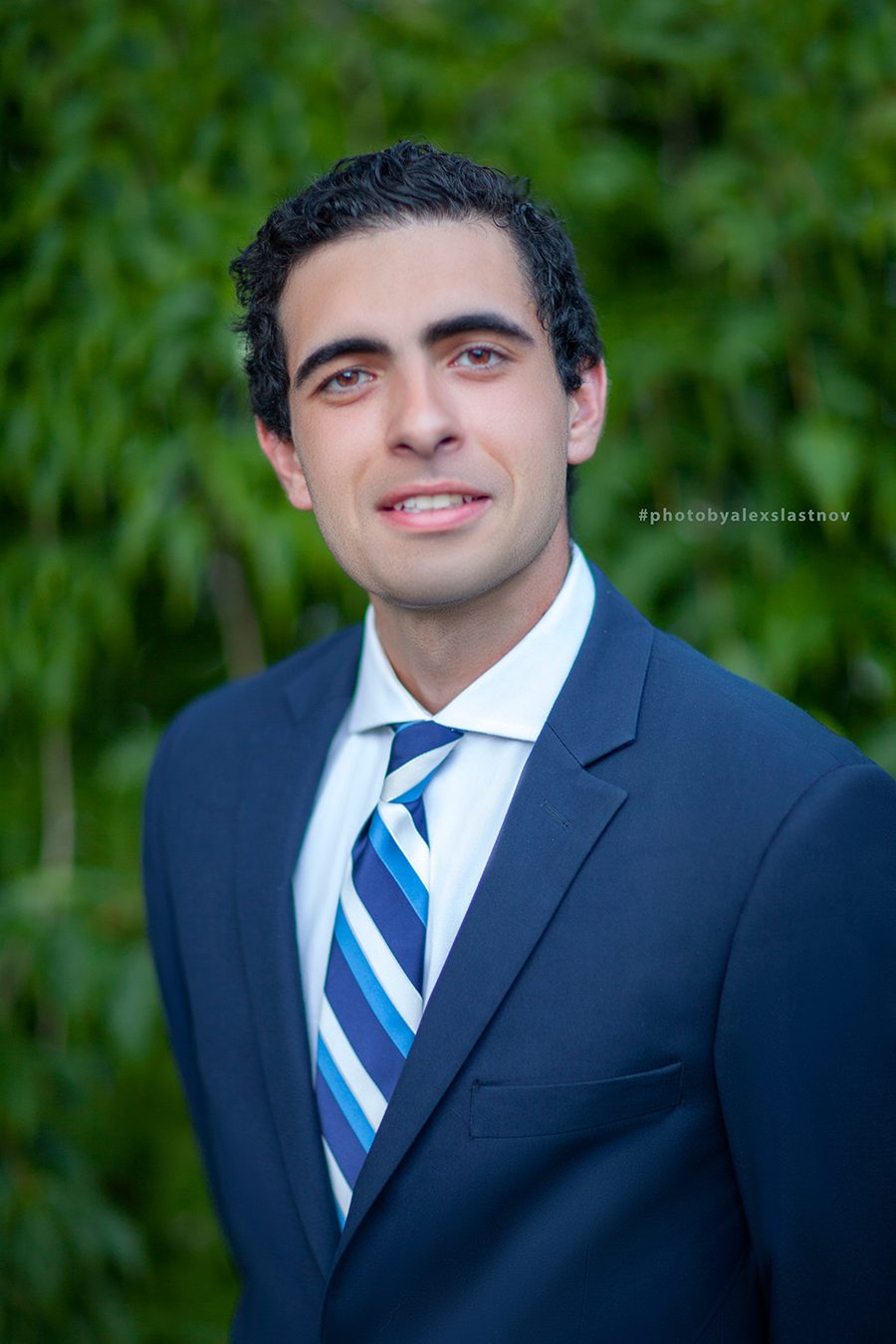 Corporate headshot of a man in a navy suit and striped tie against green foliage