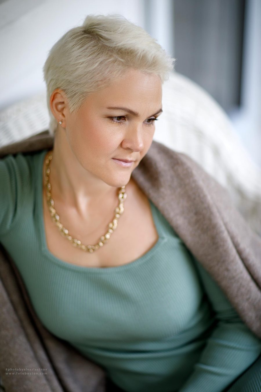 Lifestyle portrait of a woman with short blond hair in a green top and a gold necklace by a window