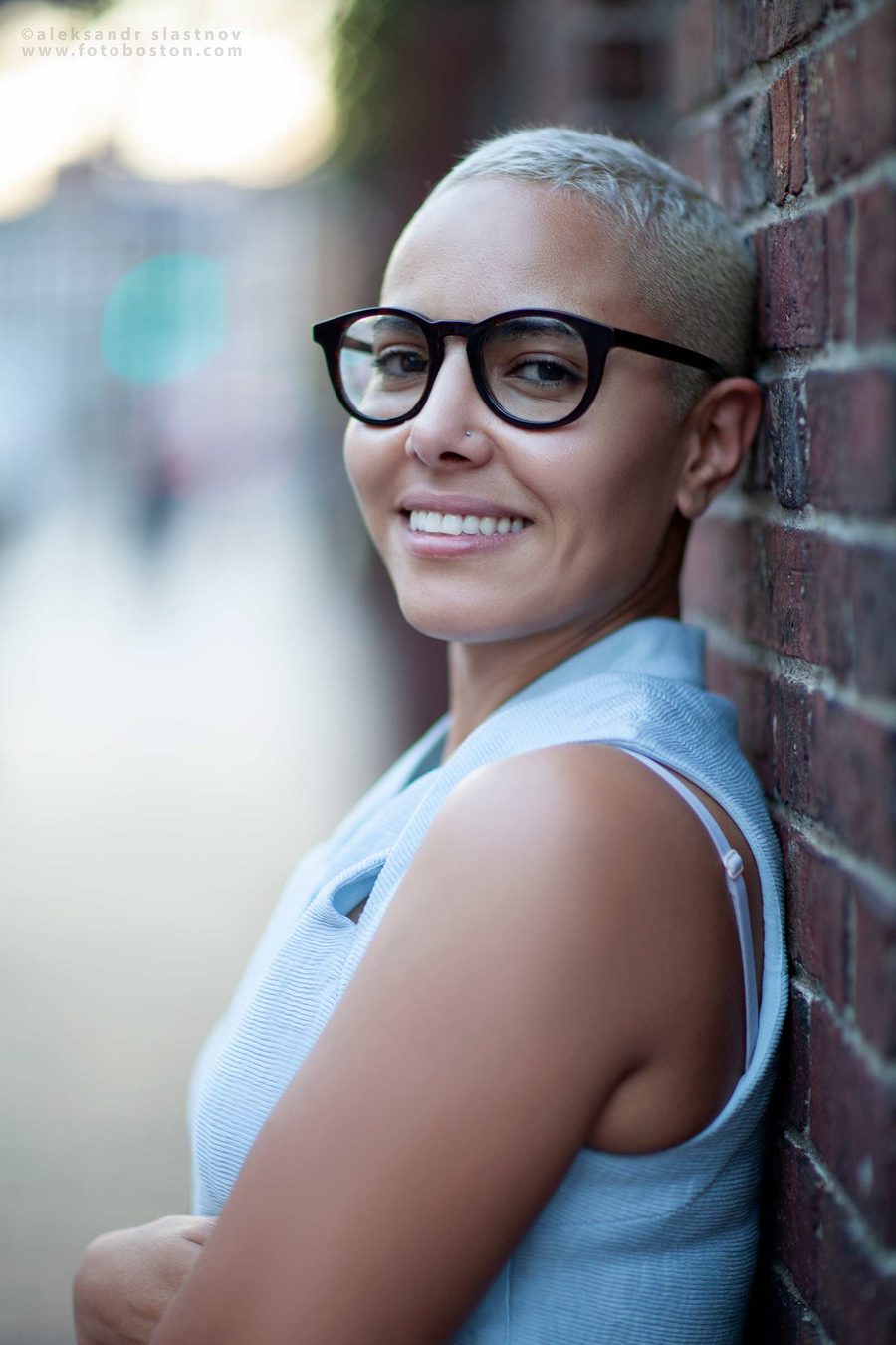 Lifestyle portrait of a smiling woman with short blond hair and round glasses leaning on a brick wall